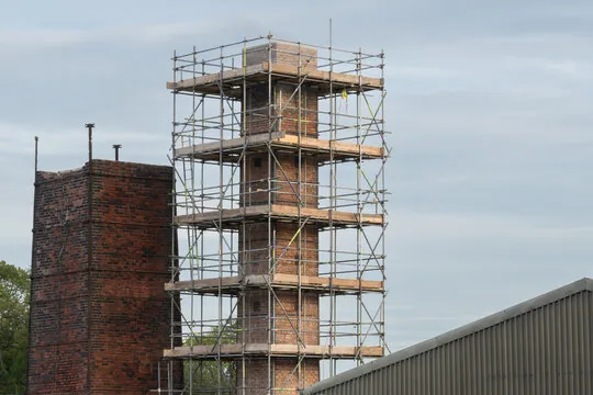 Scaffolding wrapped around a tall brick chimney next to an older chimney